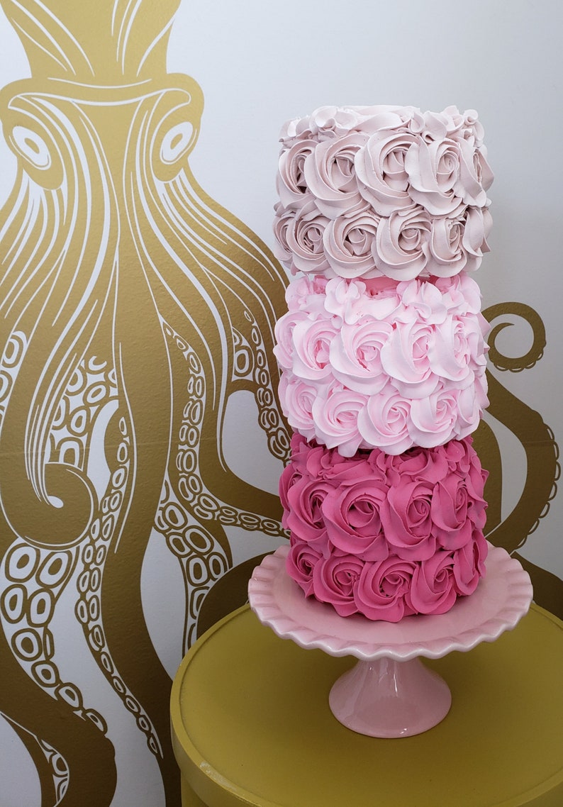 Puede incluir: Un pastel de tres pisos decorado con glaseado rosa y rosa claro, que se asemeja a rosas. El pastel est&aacute; sobre un soporte para pastel rosa con un borde festoneado. El soporte para pastel est&aacute; sobre una mesa amarilla.