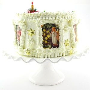K&ouml;nnte beinhalten: Ein wei&szlig;er Kuchen, verziert mit wei&szlig;em Frosting und goldenen Sternen. Der Kuchen ist mit Bildern von Engeln und Weihnachtsb&auml;umen verziert. Der Kuchen steht auf einem wei&szlig;en Kuchenst&auml;nder.