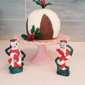 May include: Two ceramic candy cane figurines with winking faces stand on either side of a white and brown cake decorated with holly and red berries. The cake is on a pink pedestal.