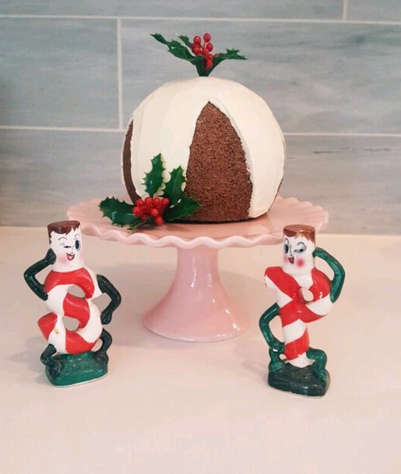 May include: Two ceramic candy cane figurines with winking faces stand on either side of a white and brown cake decorated with holly and red berries. The cake is on a pink pedestal.