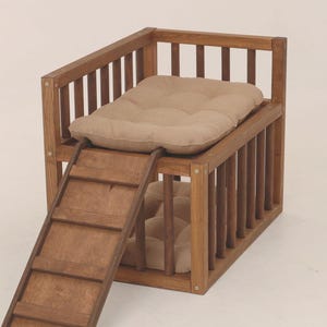 May include: A wooden pet bed with a built-in ladder. The bed has a two-tiered design with a tan cushion on each level. The frame is made of vertical wooden slats and horizontal rails. The ladder provides easy access to the upper level.