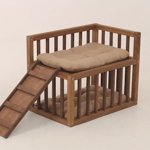 May include: A two-story wooden pet bed with a built-in ramp. The bed has a rectangular frame with vertical bars and two tan cushions. The ramp is made of wooden slats, providing easy access to the upper level. The overall design is a warm brown color.
