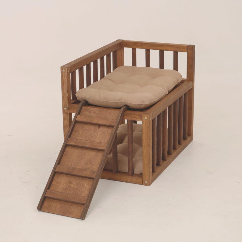 May include: A two-tiered wooden pet bed with a natural wood finish. The upper level has a beige cushion, reached by a small wooden ramp. The lower level also has a beige cushion. The bed is constructed with vertical wooden slats, offering a cosy space.