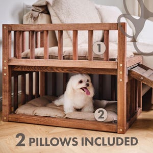 May include: A two-tier wooden dog crate with a brown finish. The crate features a slatted design and includes two beige cushions. A small white dog is inside the lower level. The text "2 PILLOWS INCLUDED" is at the bottom.