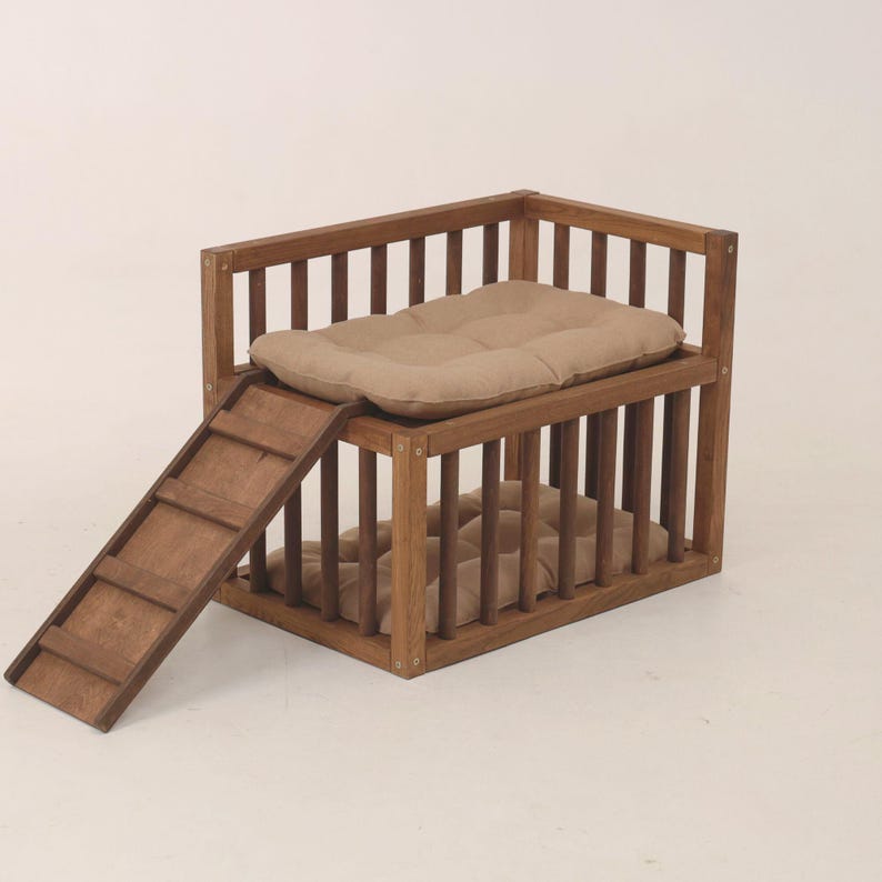 May include: A two-tier wooden pet bed with a ramp. The bed features a rectangular frame with vertical bars and two tan cushions. A wooden slatted ramp provides access to the upper level. The structure is a warm brown colour, designed for small pets.