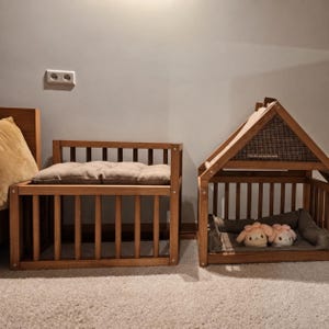 May include: A wooden pet bed frame with a beige tufted cushion and a separate wooden pet house frame with a peaked roof, featuring a grey interior cushion and two small plush toys with pink ears resting inside the structure.