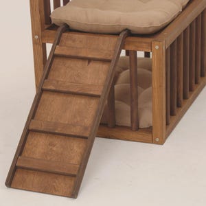 May include: A wooden pet bed with a ramp for easy access. The bed frame is made of stained wood and features vertical bars on the sides. It includes a soft, beige cushion for comfort. The ramp has four steps.