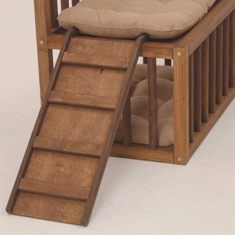 May include: A wooden pet bed with a ramp for easy access. The bed frame is made of stained wood and features vertical bars on the sides. It includes a soft, beige cushion for comfort. The ramp has four steps.