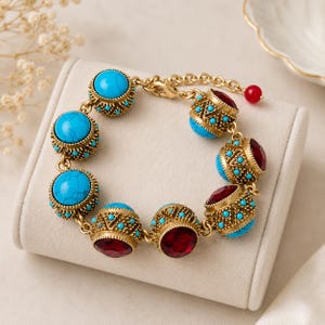 May include: A gold-tone chain bracelet featuring alternating round turquoise-colored stones and deep red faceted stones. Each stone is set within an ornate, filigree-style gold metal casing decorated with tiny turquoise beads and a lobster clasp.