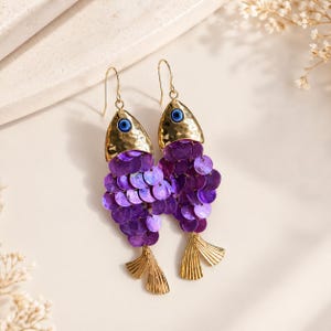 May include: A pair of unique fish-shaped earrings. The earrings feature gold-colored metal tops with blue eye details, and purple sequin bodies. The fish have gold-colored metal fins and are suspended from gold-colored hooks.