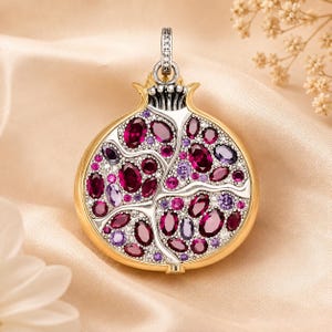 May include: A pomegranate-shaped pendant crafted from gold and silver metal, featuring an intricate arrangement of deep red and light purple oval-cut gemstones. The surface is densely encrusted with tiny clear stones, and a bail sits at the top.
