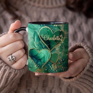May include: A black and white mug with a green and gold abstract design featuring two hearts. The larger heart has the name "Charlotte" written inside.