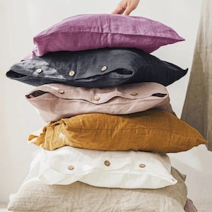 Op de afbeelding: Een stapel van zes linnen kussens in verschillende kleuren: paars, antracietgrijs, blush roze, mosterdgeel, wit en naturel beige. Elk kussen heeft een knoopsluiting. Een hand is zichtbaar, die het bovenste kussen zachtjes aanraakt.