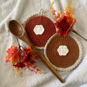 Puede incluir: Dos posavasos de ganchillo en color marrón y crema con una flor blanca de ganchillo en el centro. Los posavasos están sobre una superficie blanca y peluda con una cuchara de madera y flores de color otoñal.