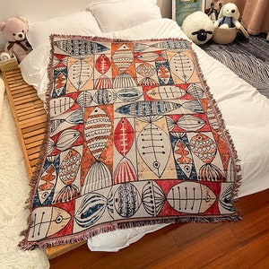 May include: A woven throw blanket with a colourful geometric pattern of stylised fish in shades of red, blue, beige, and brown. The blanket has a fringed edge.
