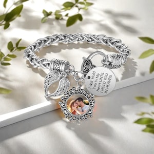 May include: Silver-toned bracelet with a woven chain design. The bracelet features a round charm with a photo and a decorative frame, a wing charm, and a circular charm with the inscription "I Will Hold You In My Heart Until I Hold You In Heaven."