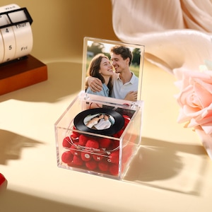 Könnte beinhalten: Eine transparente Acrylbox mit einem Foto eines Paares, einer kleinen Schallplatte mit einem Foto und roten Pompons. Die Box hat ein Foto eines Paares oben. Der Text "A Thousand Years" ist sichtbar.