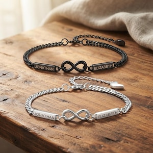 Op de afbeelding: Twee gourmetkettingarmbanden, één zwart en één zilverkleurig, elk met een centraal oneindigheidssymbool geflankeerd door twee rechthoekige metalen staafjes. De staafjes zijn gegraveerd met de namen JONATHAN en ELIZABETH. Elk armbandje heeft een hartje.