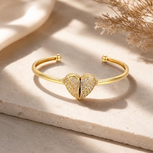 Può includere: Un bracciale rigido color oro con un ciondolo centrale a forma di cuore. Il cuore è composto da due metà, ciascuna decorata con dettagliati motivi ad ali e incastonata con piccole pietre chiare e scintillanti che riflettono la luce.