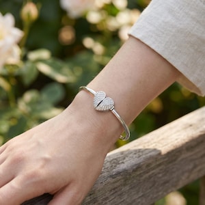 May include: A silver-toned bangle bracelet featuring a central heart-shaped charm composed of two halves. The heart is encrusted with numerous small, clear, sparkling stones that cover the entire surface of both segments of the heart design.