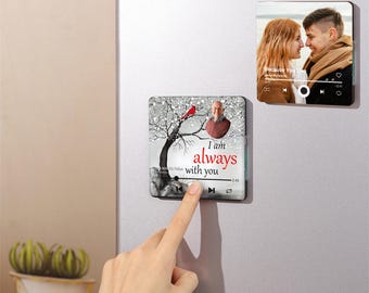 Pode incluir: Um ímã de geladeira quadrado com uma cena de inverno que apresenta uma árvore nevada, um cardeal vermelho e o rosto de um homem. O ímã tem uma interface de reprodutor de música com botões de reprodução, pausa e avanço. O texto "Estou sempre com você" está impresso no ímã.