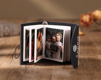 May include: Black leather photo album with the word "Daddy" embossed on the cover. The album is open to reveal photos of a family, including a father and two children. The photos are printed on white paper and have a vintage look.