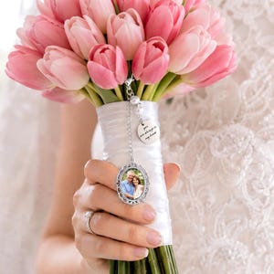 Op de afbeelding: Een boeket roze tulpen omwikkeld met wit satijnen lint, voorzien van een zilverkleurige ovale fotobedel met een portret van een koppel en een ronde metalen hanger met de tekst, You are always in my heart, bevestigd aan een zilveren ketting.