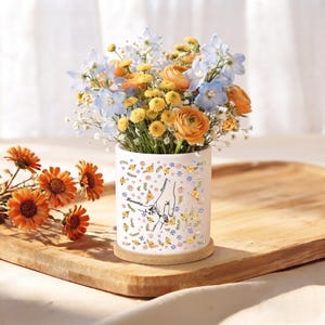 May include: A white cylindrical ceramic vase featuring a delicate floral pattern and a line art illustration of two hands holding each other. The vase is filled with a vibrant bouquet of light blue delphiniums, orange ranunculus, and yellow flowers.