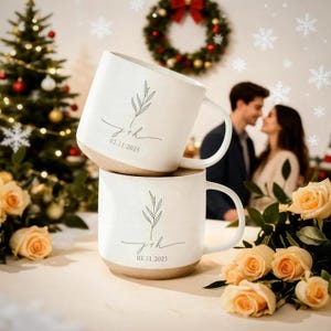 May include: Two white ceramic mugs stacked on top of each other. Each mug has a light brown base, a white handle, and a delicate green leaf design with the initials "j+h" and the date "02.11.2025". The mugs are on a table with roses and a Christmas tree in the background.