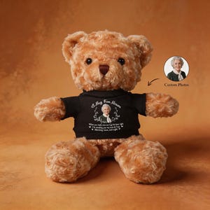 Puede incluir: Un oso de peluche marrón que lleva una camiseta negra con el texto "A Hug From Heaven" y una foto de una mujer. El oso tiene una opción de foto personalizada y un mensaje sobre amor y abrazos.