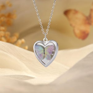 May include: Silver-toned heart-shaped pendant necklace. The pendant features a colorful butterfly design inside a heart-shaped frame. The butterfly has pastel blue, purple, and yellow wings. The necklace chain is delicate and silver.
