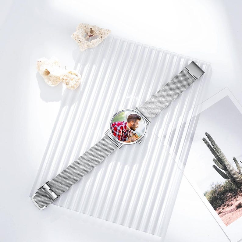 May include: A silver wristwatch with a mesh band. The watch face displays a color photograph of a person wearing a red and black plaid shirt. The watch is displayed on a white, textured surface with decorative elements.