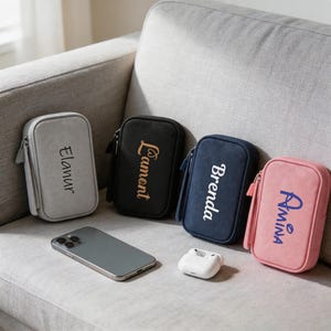 May include: Four zippered pouches in gray, black, navy, and pink, each with a name printed on the front. A smartphone and a pair of white wireless earbuds are also visible. The pouches are rectangular and have a wrist strap. The background is a gray sofa.