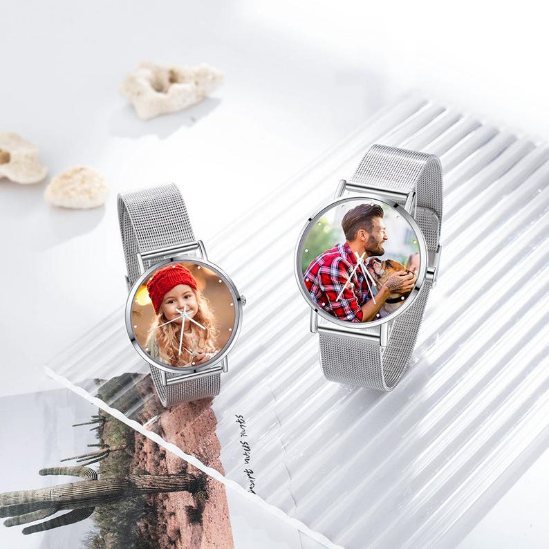May include: Two silver wristwatches with mesh bands. The watch faces display personalized photos. One watch shows a young girl in a red hat, and the other features a man with a dog. The watches are on a white surface.