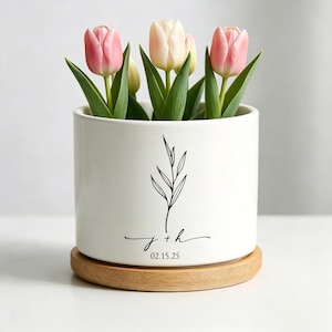 Puede incluir: Una maceta de cerámica blanca con un diseño botánico negro y la inscripción "j + h 02.15.25". La maceta contiene varios tulipanes con pétalos rosas y crema y hojas verdes. La maceta se asienta sobre una base redonda de madera marrón claro.