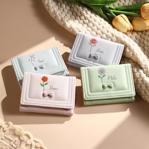 Puede incluir: Cuatro carteras pequeñas rectangulares de tres pliegues en azul claro, gris claro, rosa claro y verde claro. Cada cartera presenta un pequeño lazo en relieve, una ilustración floral y un nombre: Finn, Lean, Daine o Mila, en letra negra.