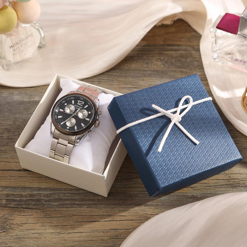May include: A silver wristwatch with a black face and multiple sub-dials rests on a white pillow inside a cream-colored box. A blue textured gift box tied with white string is next to the watch box. The watch has a stainless steel band.