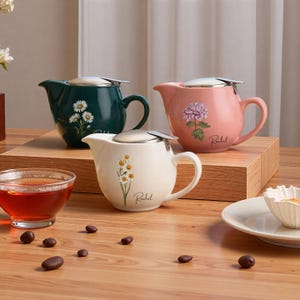 May include: Three ceramic teapots in green, pink, and cream, each with a floral design and a stainless steel infuser lid. The teapots have a curved handle and spout. The cream teapot has a daffodil design, the pink teapot has a peony design, and the green teapot has a daisy design.