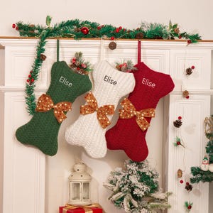 Puede incluir: Tres calcetines navideños en verde, blanco y rojo, cada uno con un lazo y el nombre "Elise". Los calcetines cuelgan de una repisa blanca decorada con vegetación y adornos festivos, creando una escena navideña.