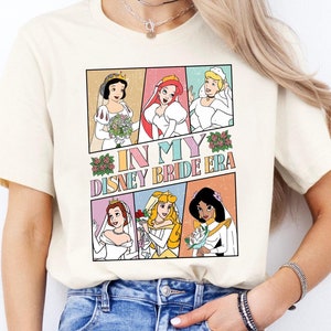 May include: Cream-colored t-shirt featuring a colorful graphic of Disney princesses in wedding attire. The graphic includes the text "IN MY DISNEY BRIDE ERA" in a retro comic book style.