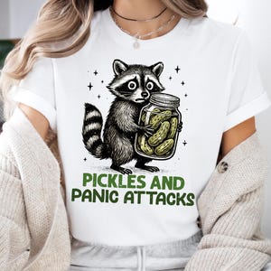 May include: White t-shirt featuring a black and white raccoon holding a jar of pickles. The raccoon has a worried expression. The text "PICKLES AND PANIC ATTACKS" is printed below the raccoon in green.