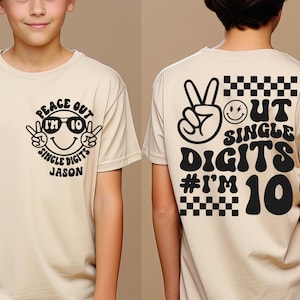 May include: A beige t-shirt with black graphics. The front features a smiling face wearing sunglasses with the text "PEACE OUT I'M 10 SINGLE DIGITS JASON". The back has a peace sign, smiley face, and text "OUT SINGLE DIGITS #I'M 10".