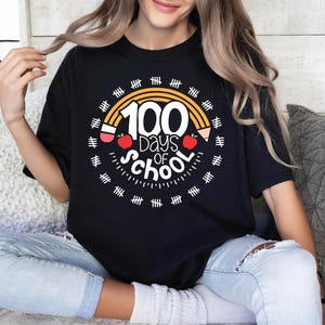 Puede incluir: Camiseta negra con un gráfico blanco de un arcoíris con el texto "100 días de escuela" y un lápiz y una manzana.