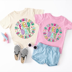 May include: Two children's t-shirts, one cream and one pink, with the words "FIELD DAY" in a colorful design. Accessories include blue denim shorts, checkered trainers, and sunglasses. Crayons are also visible.