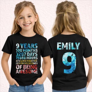 Op de afbeelding: Zwart T-shirt met de tekst "9 YEARS, 108 MONTHS, 3287 DAYS, 78888 HOURS, 4733280 MINUTES, 283996600 SECONDS OF BEING AWESOME" op de voorkant. De achterkant toont "EMILY" en het cijfer "9".