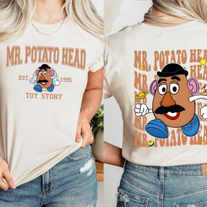 Può includere: Una maglietta beige con una grafica marrone di Mr. Potato Head dal film Toy Story. Il testo sulla maglietta recita "Mr. Potato Head, Est. 1995, Toy Story". La parte posteriore della maglietta ha un motivo ripetuto del testo "Mr. Potato Head" in un font marrone retrò.