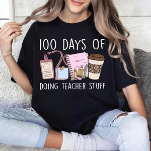 Puede incluir: Camiseta negra con el texto "100 DAYS OF DOING TEACHER STUFF". El diseño incluye una insignia, un bloc de notas, lápices, notas adhesivas y una taza de café con estampado de leopardo.