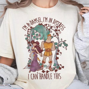 May include: A white t-shirt with a vintage-style illustration of a cartoon couple. The text on the shirt reads "I'm a-damsel, I'm in distress I can't handle this".