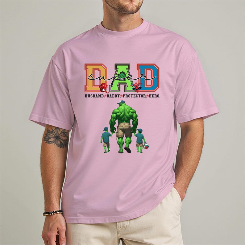A man wearing a pink t-shirt with a graphic design featuring a muscular green character and text that reads "SUPER DAD" and "Husband, Daddy, Protector, Hero."