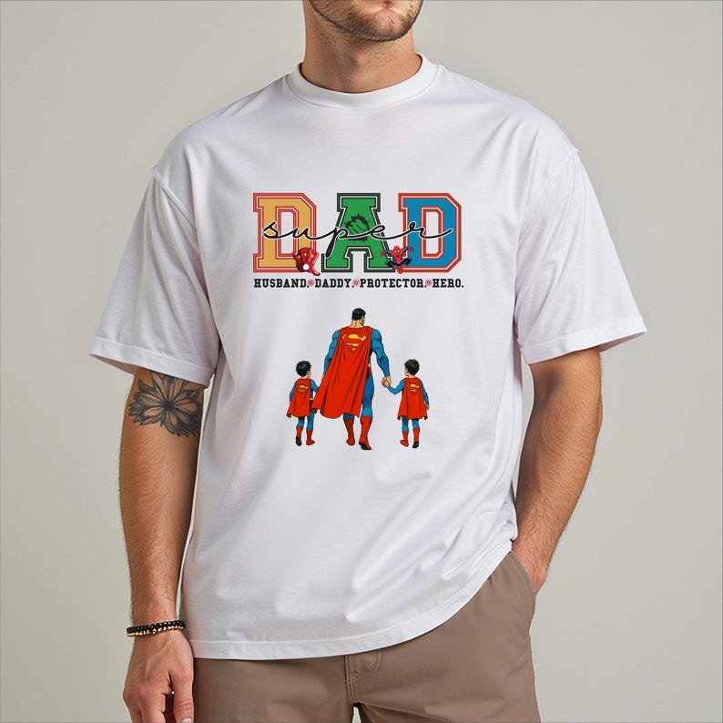 A white t-shirt with a graphic design featuring a man in a superhero costume holding three children, along with the text "DAD" and "Husband-Daddy-Protector-Heroes" printed on it.